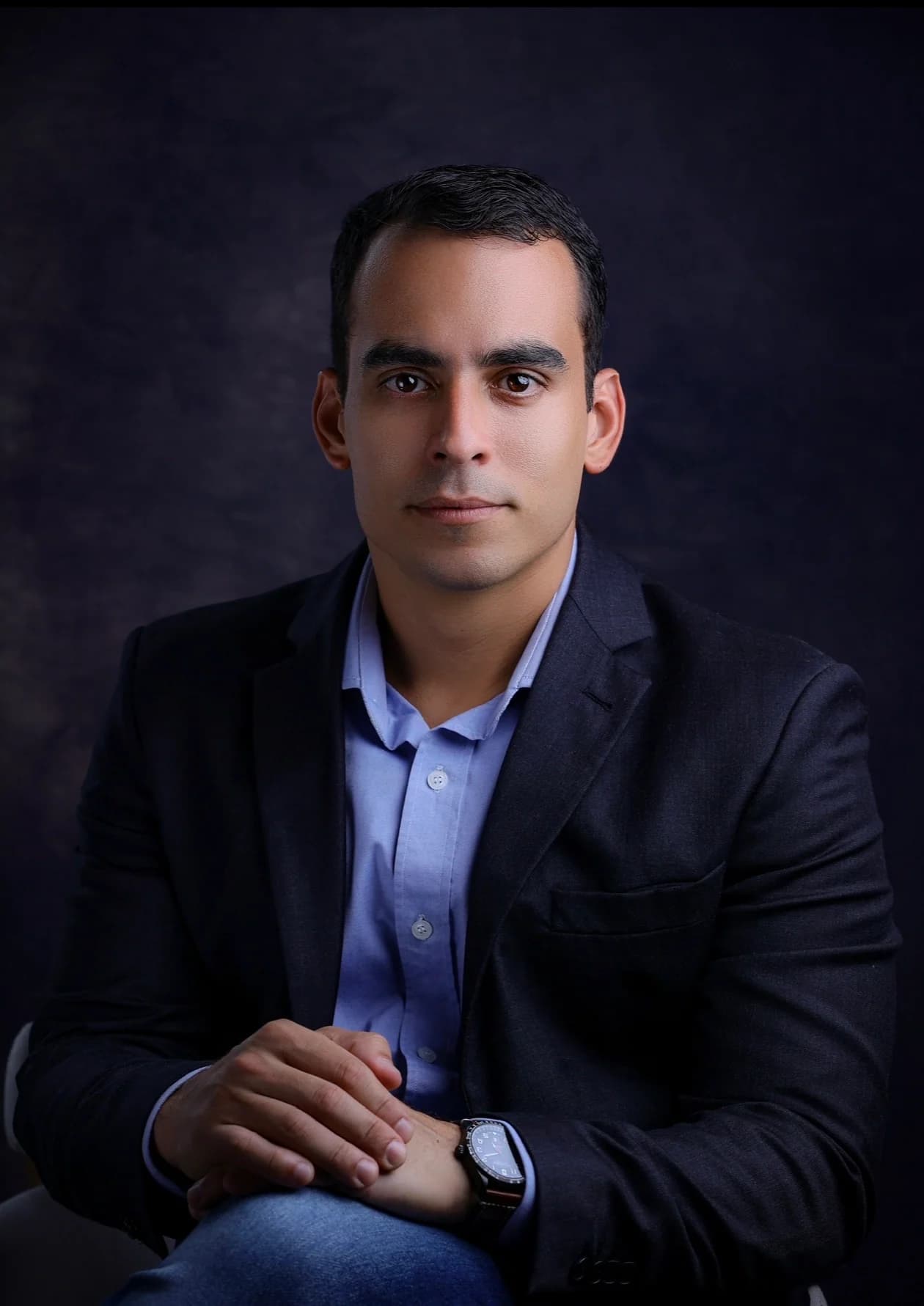 Vitor Novaes sentado, de blazer escuro, em retrato de estúdio